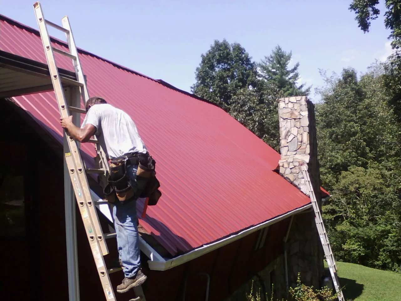 Roofer on a ladder beside a red metal roof and stone chimney — Soffit & Fascia Repair by Roofing Brothers in Woodbury